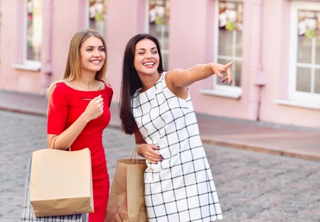 Happy young women with shopping bags pointing finger somewhere on old city background. Sale, consumerism and people conceptの写真素材