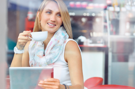 Trendy young woman in cafe with cup of coffee and touchscreen tablet while siting in cafeの写真素材
