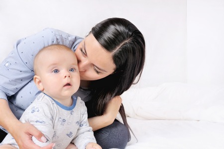 Mother and child on a white bed. Mom and baby boy playing in sunny bedroom. Family having fun togetherの写真素材