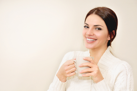 A beautiful smiling young woman with long hair and brown eyes drinking coffee or tea from a cup. Beautiful adult girl hods in hands mug with hot drinkの写真素材
