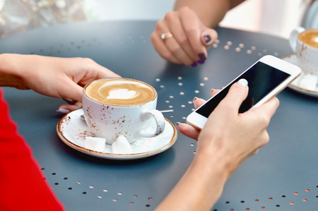 Close up view of two coffee cups and hands of young women with smartphone at outdoor cafe. Lifestyle, friendship and technology conceptの写真素材