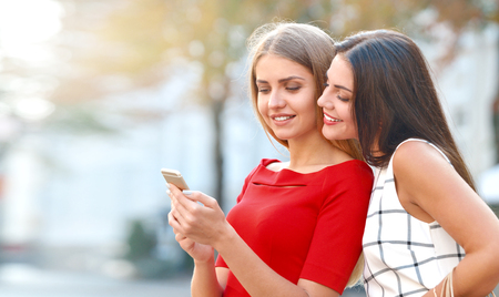 Woman shows on a mobile phone something to his girlfriend. Happy young women with smartphone on city streetの写真素材