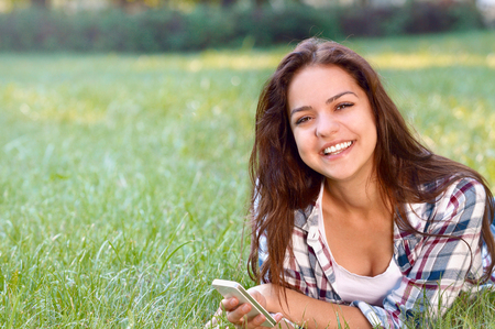 Happy brunette woman lying on grass in park with smartphone and looking at the camera. Young student girl lying with smartphone in her hands. Communication and social network concept.の写真素材