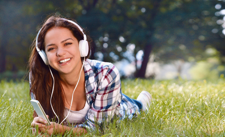 Portrait of pretty young girl listening music lying at the grass. Cute teenage girl relaxing in parkの写真素材