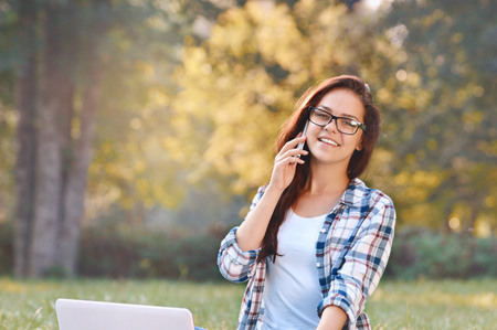 Student girl working on laptop, sitting on grass in park. Student studying outdoors. Freelance job, online shopping, distance learning conceptの写真素材