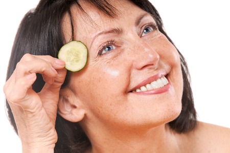 Cheerful mature woman holding pieces of cucumber near of her eyes. Natural spa treatment. Isolated on white backgroundの写真素材