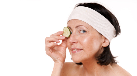 Cheerful mature woman holding piece of cucumber in front of her eye. Natural spa treatment. Isolated on white backgroundの写真素材