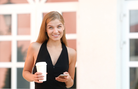A businesswoman checking email via mobile phone and holding a coffee cup against urban sceneの写真素材