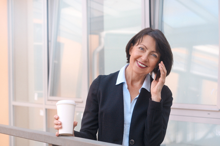 Mature businesswoman having a break in front of an office building, talking on the phone and drinking coffee. Work anywhere concept.の写真素材