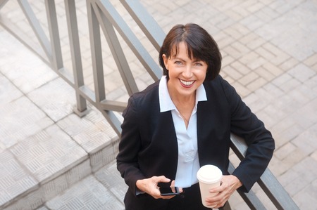 Elderly business woman with coffee using cell phone, outdoor. Work anywhere concept.の写真素材