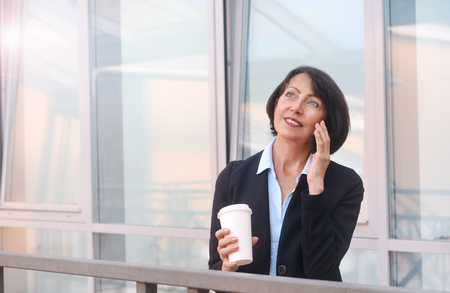 Mature businesswoman having a break in front of an office building, talking on the phone and drinking coffee. Work anywhere concept.の写真素材