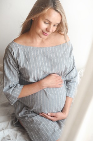 Happy pregnant woman sit on a bed at home and looking to the camera. Happy motherhood concept. Verticalの写真素材