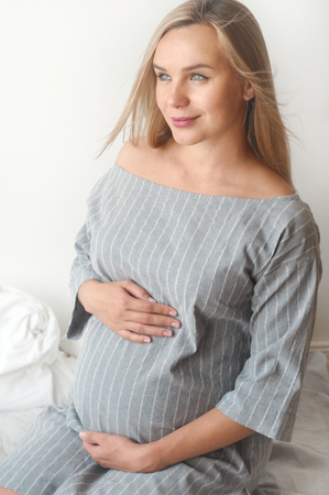 Happy pregnant woman sit on a bed at home and looking to the camera. Happy motherhood concept. Verticalの写真素材