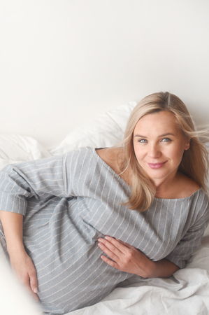 Beauty pregnant woman lie on a bed at home looking at camera. Happy motherhood concept. Verticalの写真素材
