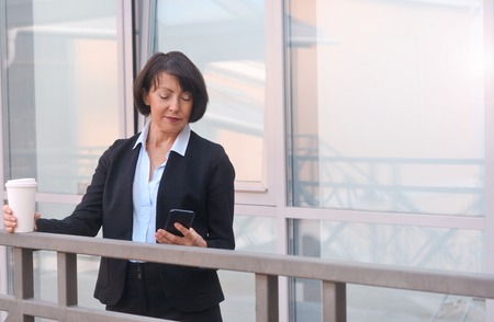 Mature businesswoman with a smartphone and cup of coffee is standing on city street next to business center. Work anywhere concept.の写真素材