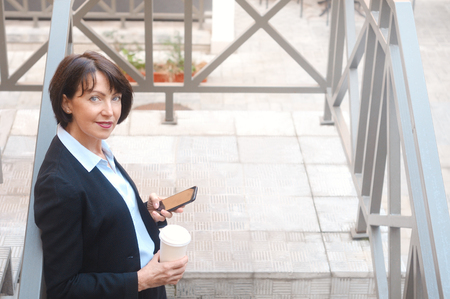 Elderly business woman with coffee using cell phone, outdoor. Work anywhere concept.の写真素材