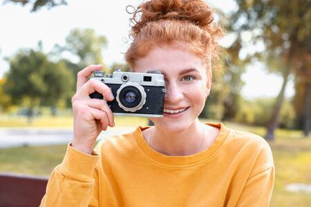 Smiling happy red hair student girl taking picture with old vintage camera outside in autumn park. Study students conceprt.の写真素材