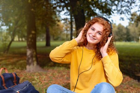 Smiling happy red hair student girl in headphones listening music outside in autumn park. Study students conceprt.の写真素材