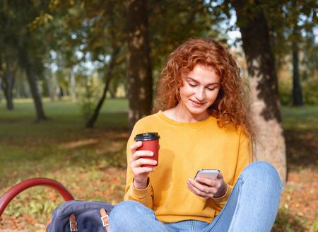 Smiling happy red hair student girl having a break with coffee outside in autumn park. Study students conceprt.の写真素材