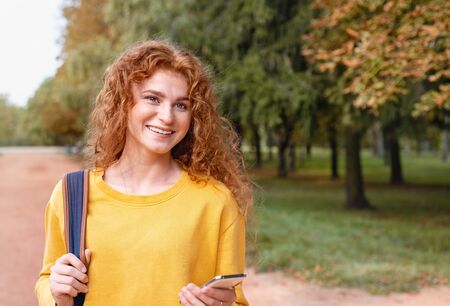 Smiling happy red hair student girl with phone outside in autumn park. Study students conceprt.の写真素材