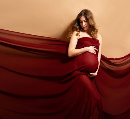 Pregnant woman with red fabric flying on wind on brown background. Motherhood conceptの写真素材