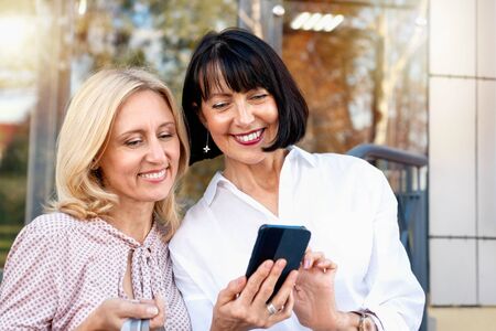 Two mature women looking at the phone and having fun while doing shopping. Pastime concept of mature peopleの写真素材