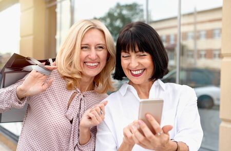 Two mature women looking at the phone and having fun while doing shopping. Pastime concept of mature peopleの写真素材