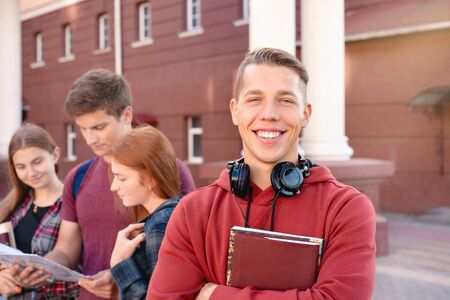 Portrait of happy smiling teenage boy with books and headphones against a group of students near university. Education conceptの写真素材