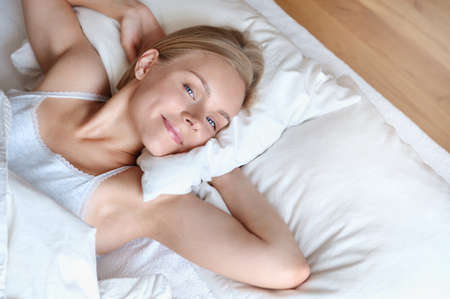 Beautiful young blonde woman waking up and stretching in the morning in a white bed. Self care lifestyle conceptの写真素材