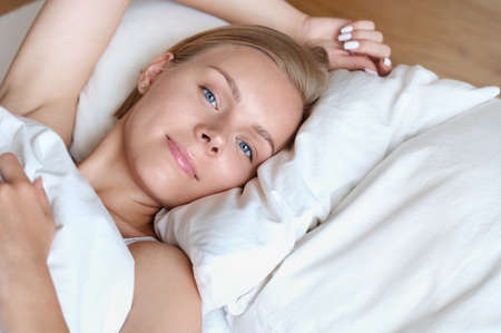 Beautiful young blonde woman waking up in the morning in a white bed. Self care lifestyle conceptの写真素材