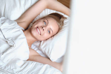Beautiful young blonde woman waking up and looking out the window in the morning in a white bed. Self care lifestyle conceptの写真素材