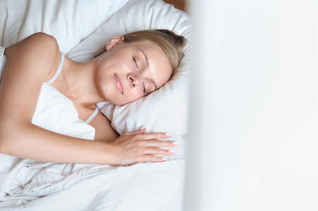 Beautiful young blonde woman sleeping near the window in a white bed. Self care lifestyle conceptの写真素材