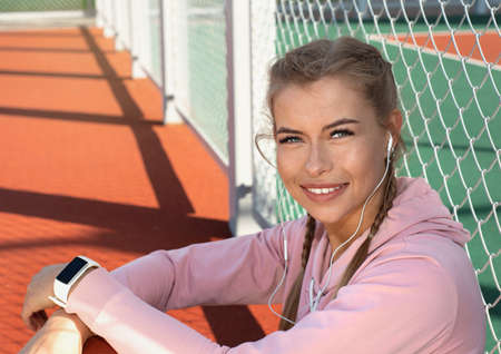 Portrait of confident smiling sporty woman wearing pink hoodie with white fitness bracelet and headphones on outdoor court. Healthy lifestyle concept.の写真素材