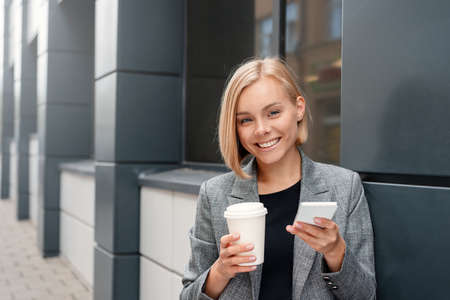 A beautiful blonde business woman with the phone with cup of coffee at the business center outdoors. Working lifestyle conceptの写真素材