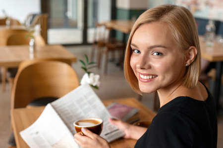 Modern beautiful blond woman reading newspaper while drinking coffee at coffee shop interior, internet distance work, internet distance work. Freelance working conceptの写真素材