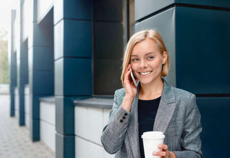 A beautiful blonde business woman on the phone with cup of coffee at the business center outdoors. Working lifestyle conceptの写真素材
