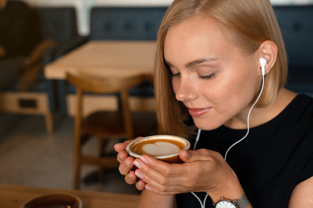 Young blonde woman listening podcast while drinking coffee at the coffe shop. Productive lifestyle concept.の写真素材