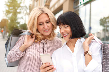 Two mature women looking at the phone and having fun while doing shopping. Pastime concept of mature peopleの写真素材