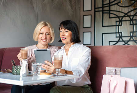 Two mature women chatting and using the tablet device while having lunch in cafe. Leisure concept of mature peopleの写真素材