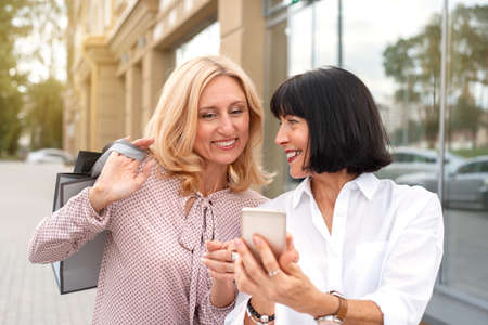 Two mature women looking at the phone and having fun while doing shopping. Pastime concept of mature peopleの写真素材