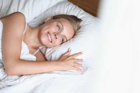 Beautiful young blonde woman waking up in the morning in a white bed. Self care lifestyle conceptの写真素材