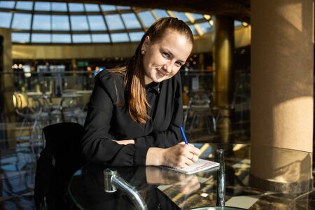 Business girl in black sits at a table, looks at the camera with a smile and fills out a documentの写真素材