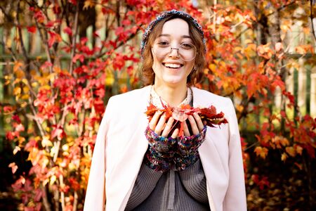 Beautiful slender girl with glasses standing on a background of red autumn leaves. Warmly dressed woman in autumn smilingの写真素材