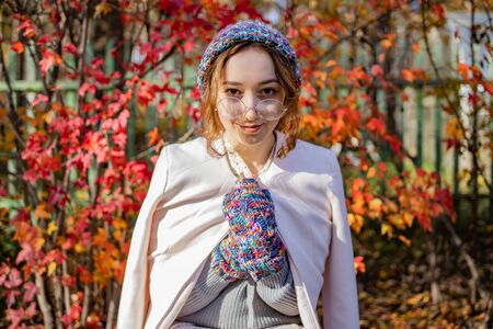 Beautiful girl in glasses stands on a background of red autumn leaves. Woman wrapped herself in warm clothes in autumnの写真素材