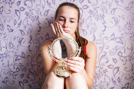 young girl looks thoughtfully into a small vintage mirror on a background of a vintage wall with cream on her fingerの写真素材
