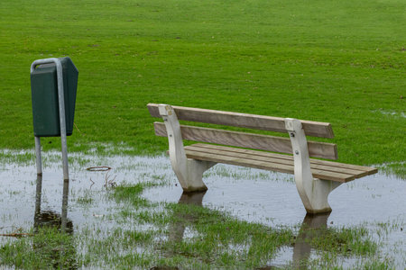 A public bench and trash can in the grass surrounded by water from heavy rain in a park.の写真素材