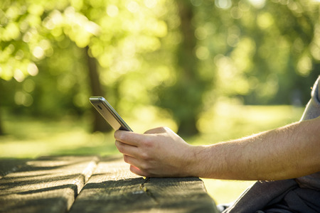 Man relaxing in park and using mobile smart phone for social media leisure. Closeup on hand holding cell phone and texting. Green nature environmet bokeh background. Wireless technology and internet business concept.の写真素材