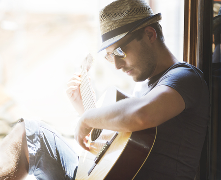 Portrait of young stylish guy with hat and sunglasses plays guitar at home. Creative man have leisure time with music at summer holiday. Hipster lifestyle concept.の写真素材