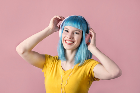 Beautiful smiling young woman with blue color dyed hair with pink headphones listening to music and dance in front of pastel studio background.の写真素材