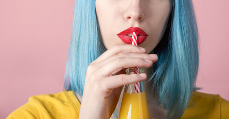 Closeup studio portrait of hipster young woman with red lipstick and blue dyed hair drinking orange juice from retro bottle with straw. Closeup of female mouth drinking summer cocktail. Trendy pastel color background. の写真素材